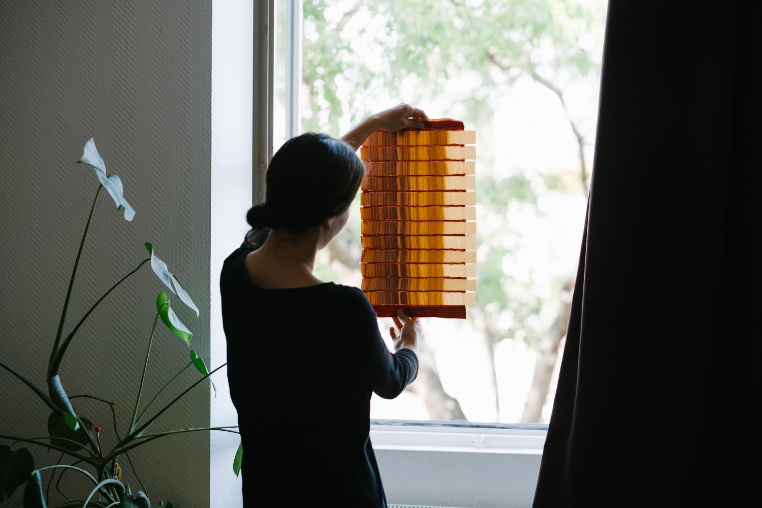 L'artiste de dos, elle tient une de ses pièces devant la fenetre pour montrer la transparence des matières.